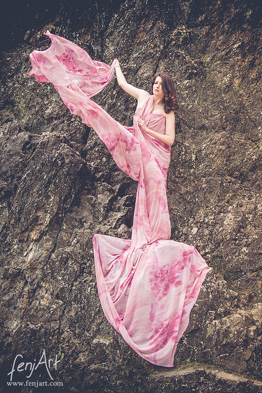 fenjArt Portraitfotografie - braunhaarige frau steht in einem rosafarbenem kleid mit wallendem tuch in einer felswand