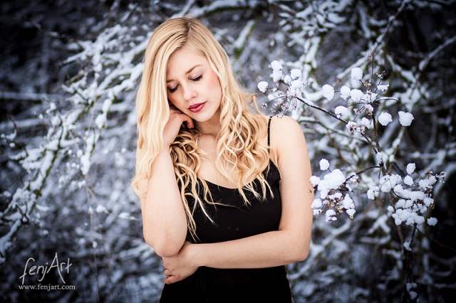 Fotoshooting mit fenjArt Fotografie blonde frau steht in schwarzem kleid vor schneebedecktem gebuesch in frankfurt und stuetzt vertraeumt das gesicht in die hand